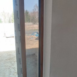 M-BUD Sp. z o.o. - Nowe okno PCV z brązową ramą i białym profilem montażowym Wikęd, widok z wnętrza na plac budowy.