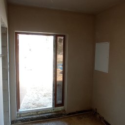 M-BUD Sp. z o.o. - Surowe wnętrze pokoju z oknem balkonowym, ściany po nałożeniu tynku, podłoga w trakcie przygotowania pod posadzkę, widoczna instalacja elektryczna przy podłodze.