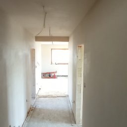 M-BUD Sp. z o.o. - Korytarz w trakcie prac wykończeniowych, ściany pokryte świeżo nałożoną, jeszcze wilgotną gładzią, widoczna drabina w oddali i wystające kable z sufitu.