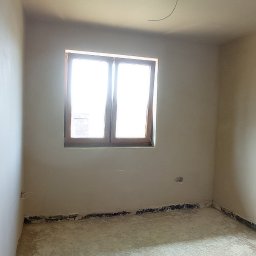 M-BUD Sp. z o.o. - Wnętrze pokoju po nałożeniu gładzi, widoczne ślady po taśmie malarskiej przy podłodze, surowe wykończenie narożnika ściany, okno z drewnianą ramą wpuszczające jasne światło.