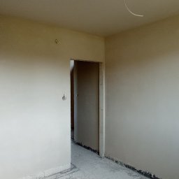 M-BUD Sp. z o.o. - Wnętrze pokoju po nałożeniu tynku na ściany, widoczne surowe wykończenie podłogi i niezabezpieczone kable wystające z sufitu.