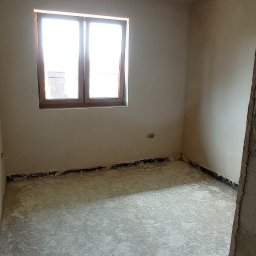 M-BUD Sp. z o.o. - Surowe wnętrze pokoju po nałożeniu tynku, widoczne ślady po pracach na podłodze, wystający przewód z sufitu i okno z drewnianą ramą.