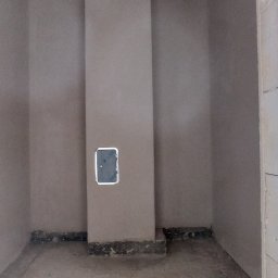 M-BUD Sp. z o.o. - Surowe wnętrze pomieszczenia po tynkowaniu maszynowym, widoczny pion kanalizacyjny z rewizją, sufit z wystającym przewodem elektrycznym, ściany w kolorze beżowym, podłoga betonowa.