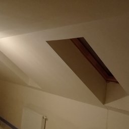 M-BUD Sp. z o.o. - Poddasze po remoncie, widoczne okna dachowe, świeżo pomalowane ściany w odcieniach beżu, surowa żarówka na kablu, zabezpieczona folia malarska na podłodze.