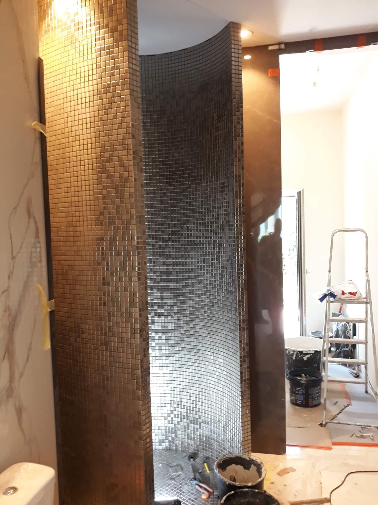 Półokrągła kabina prysznicowa wyłożona mozaiką w odcieniach brązu i szarości w trakcie montażu, widoczne narzędzia i wiaderka z zaprawą, drabina malarska w tle.