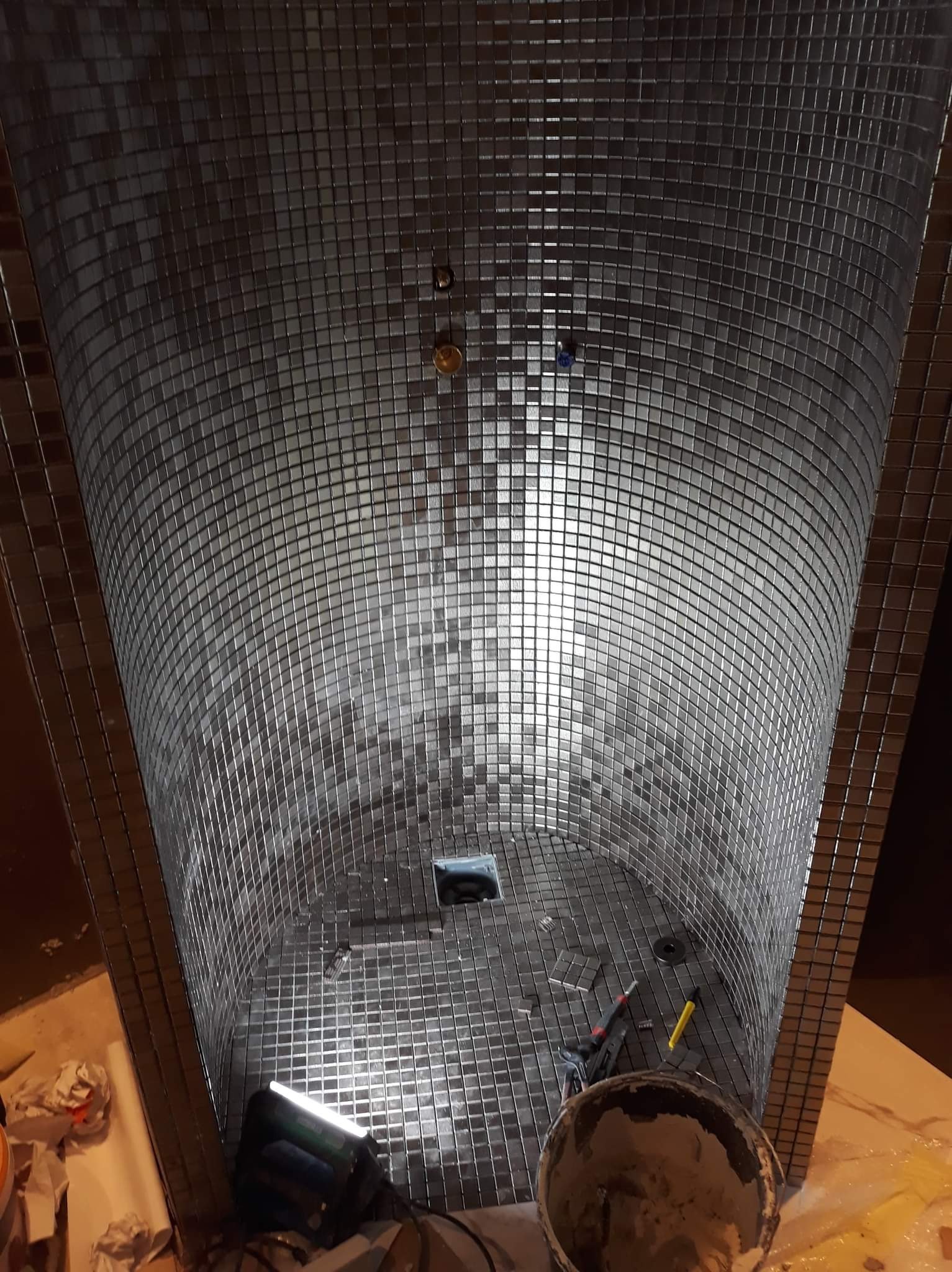 Wykończenie kabiny prysznicowej mozaiką z drobnych, srebrnych płytek; widoczny odpływ liniowy, narzędzia glazurnicze i wiadro z zaprawą.