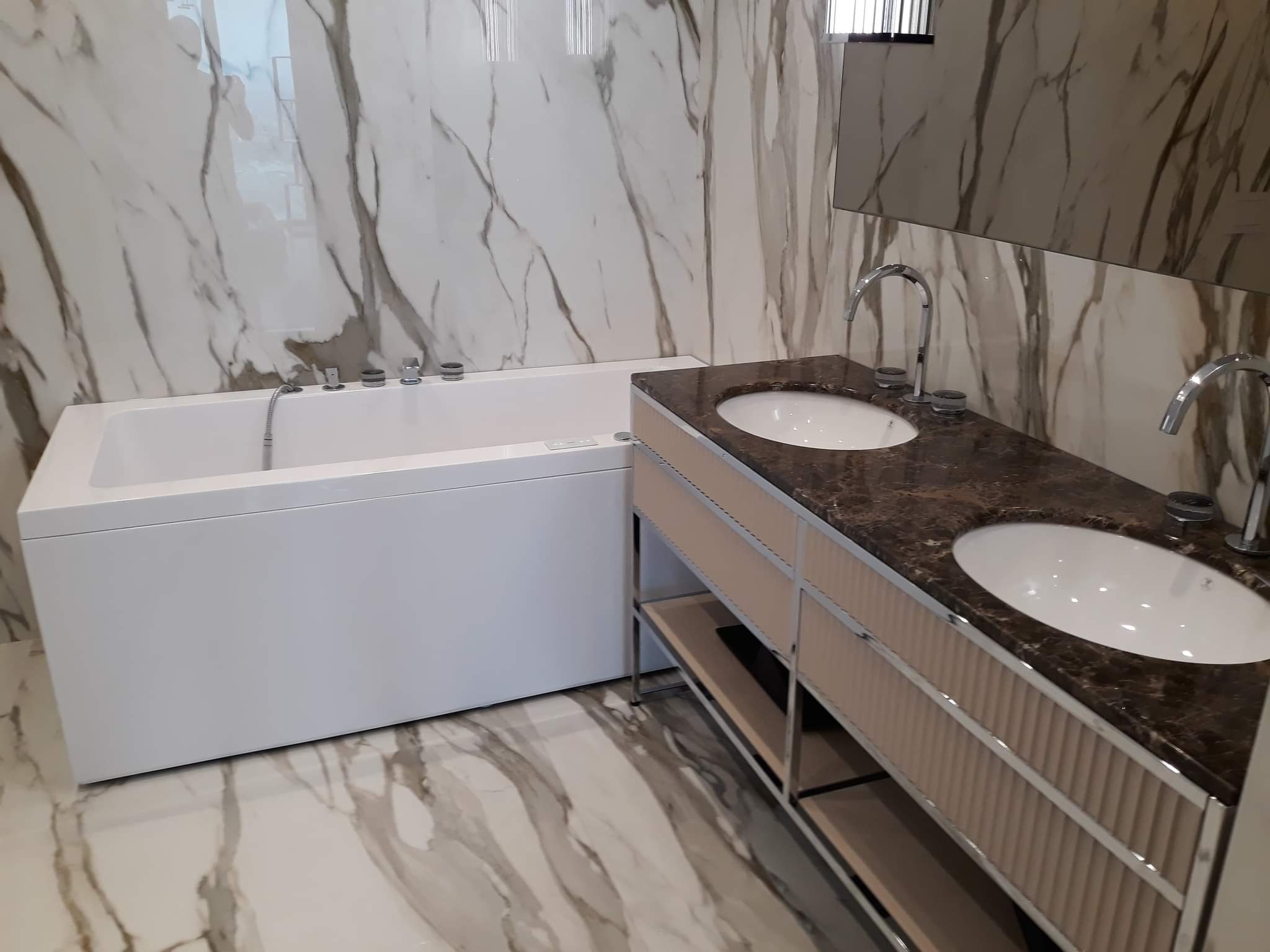 Nowoczesna łazienka z białą wanną, umywalką nablatową na brązowym blacie z szafką w beżowe pionowe paski i marmurowymi płytkami na ścianach i podłodze.