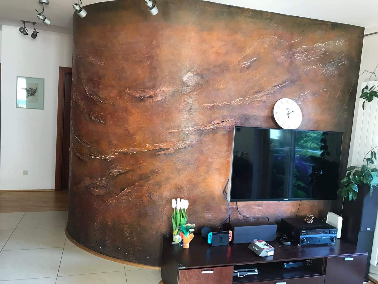 Dekoracyjna ściana w salonie z efektem rdzy i nieregularną fakturą, oświetlona punktowymi lampami sufitowymi, z widocznym telewizorem i elementami wyposażenia wnętrza.