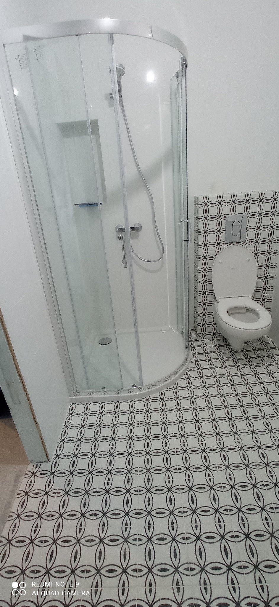 Nowoczesna łazienka z kabiną prysznicową z zaokrąglonym szkłem, toaletą z deską i wzorzystymi płytkami podłogowymi oraz na ścianie za toaletą.