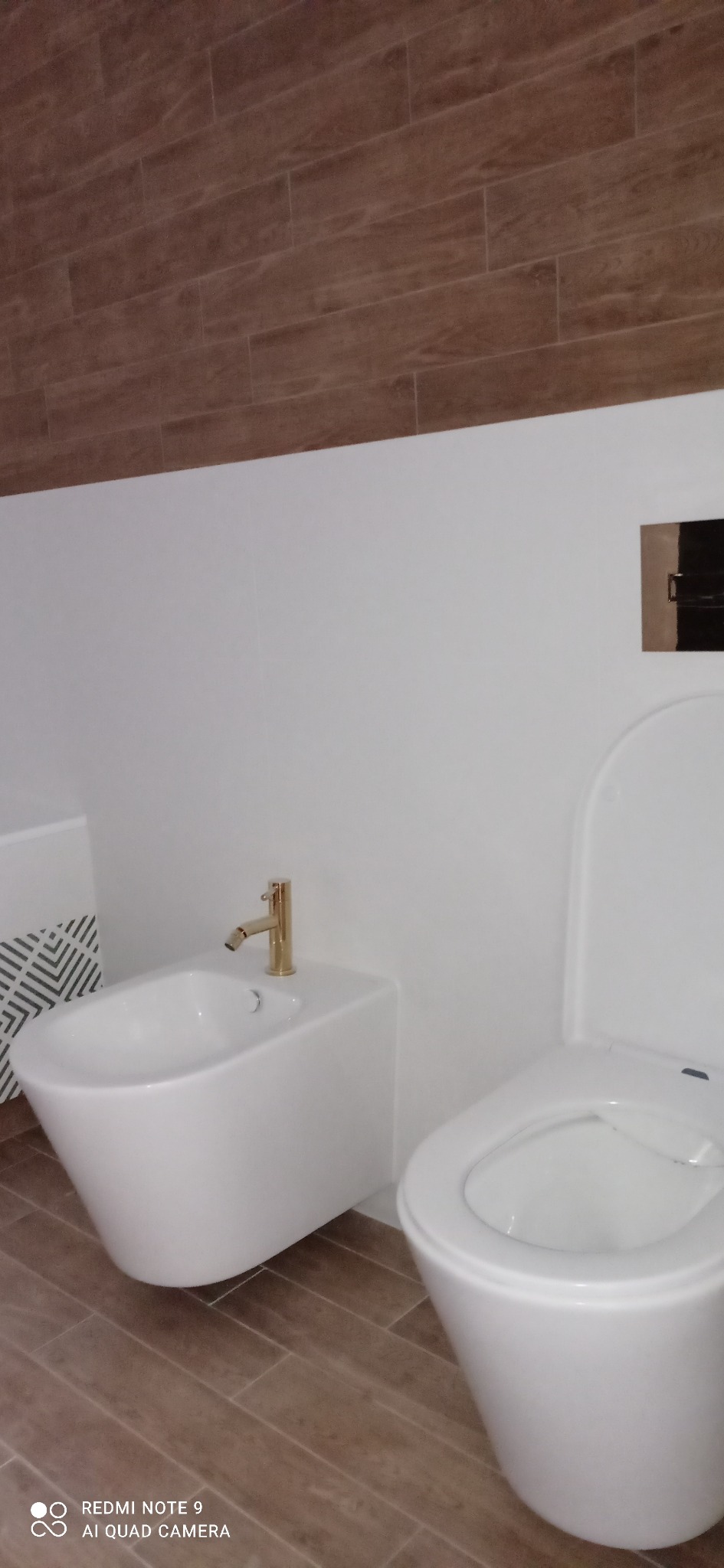 Nowoczesna łazienka z białym bidetem z pozłacaną armaturą i podwieszaną toaletą, wykończona płytkami imitującymi drewno na podłodze i ścianie.