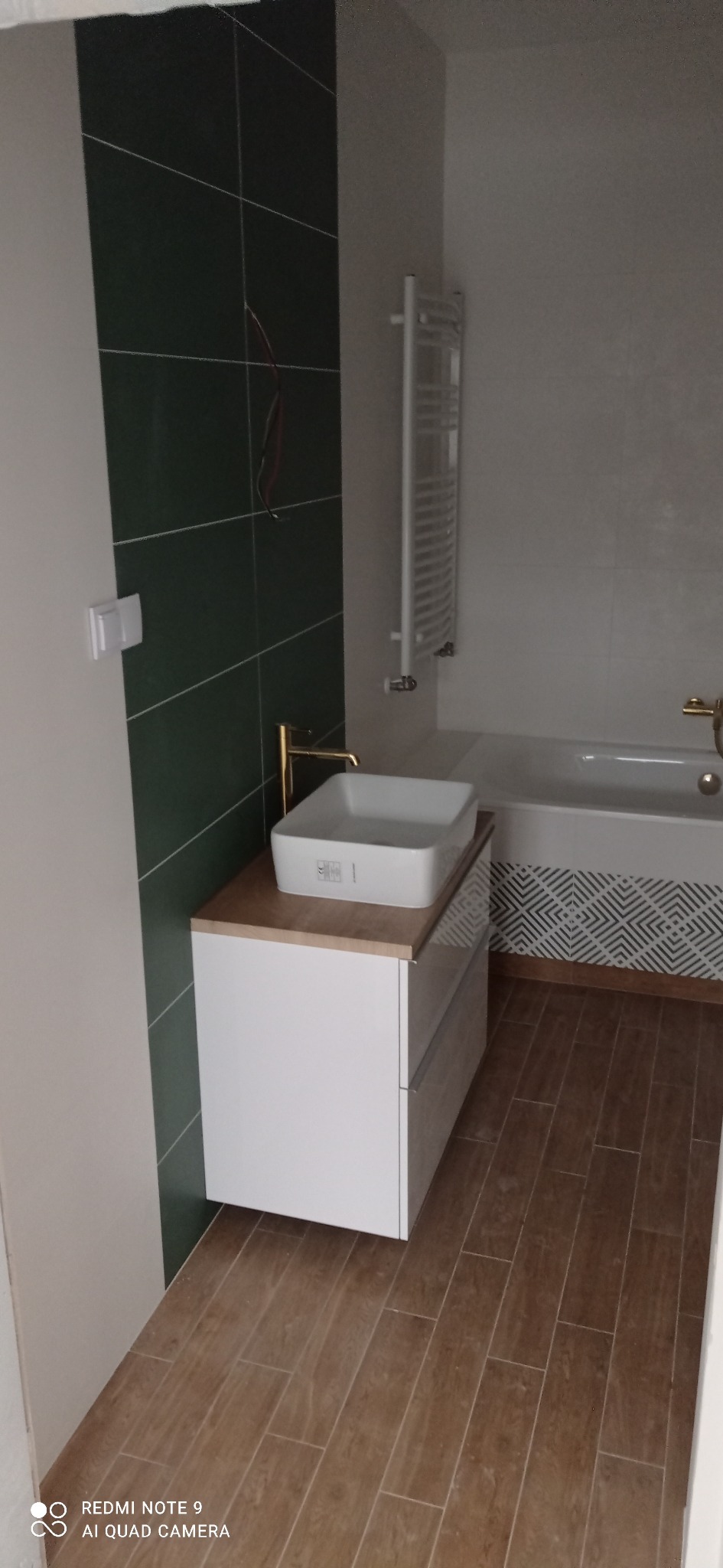 Nowoczesna łazienka z zielonymi płytkami, białą umywalką nablatową na szafce z drewnianym blatem, złotą baterią, grzejnikiem drabinkowym i wanną z geometrycznym wzorem na obudowie, brązowe płytki...