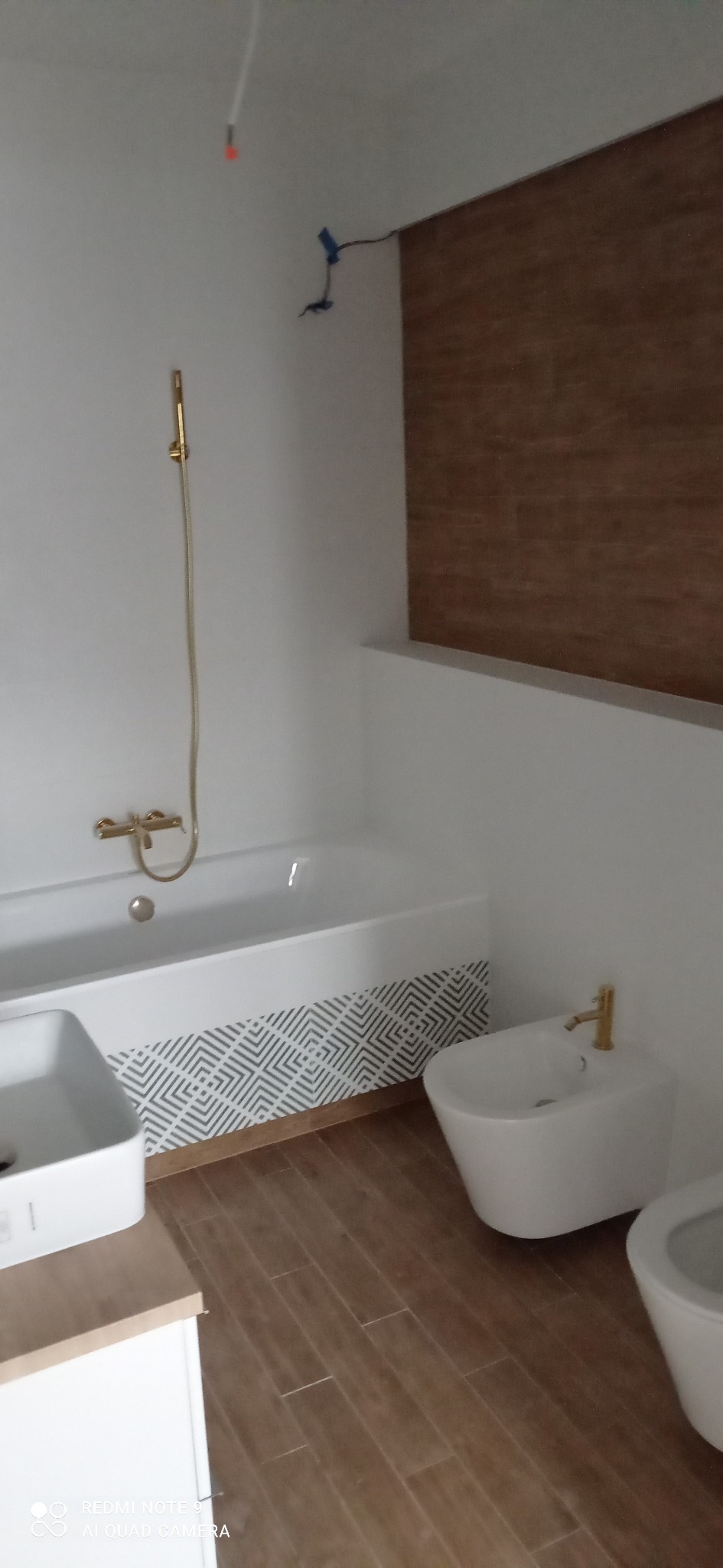 Nowoczesna łazienka z białymi sanitariatami, złotą armaturą, drewnopodobnymi płytkami podłogowymi i dekoracyjnym pasem płytek na obudowie wanny.