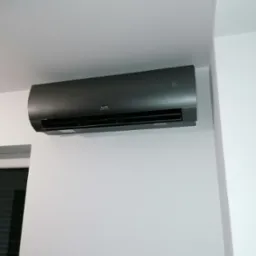 Czarny klimatyzator ścienny marki CH, zamontowany na białej ścianie w minimalistycznym wnętrzu. Widoczny fragment okna w lewym dolnym rogu.