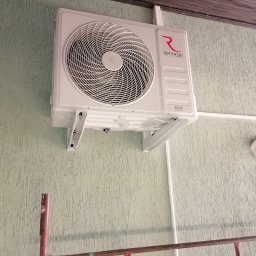 Eko-Mont- Klimatyzacja Wentylacja - Biała jednostka klimatyzacji Rotenso Roni Series zamontowana na zielonej, teksturowanej ścianie, widoczne metalowe wsporniki i fragment rusztowania malowanego na czerwono.