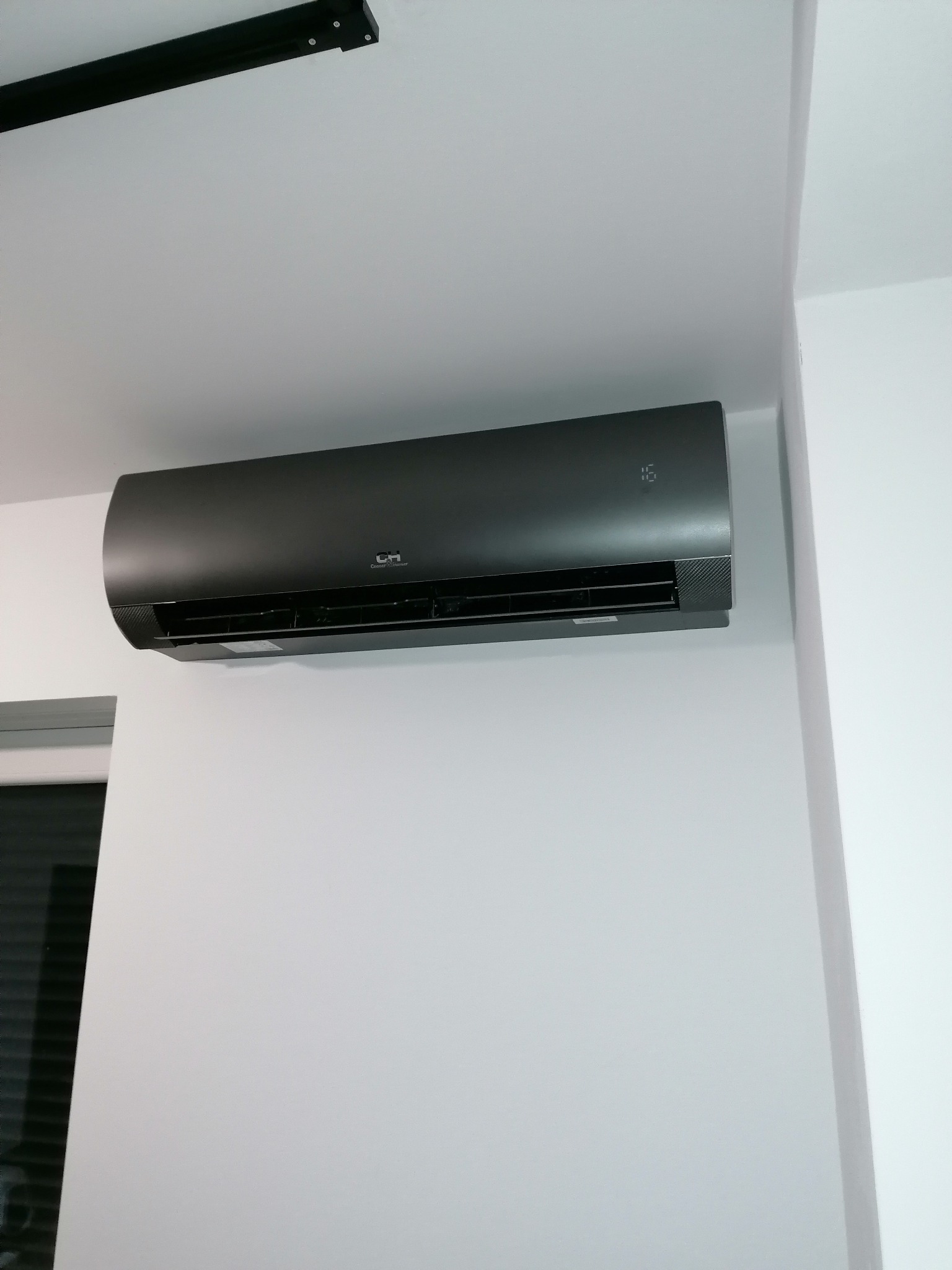 Czarny klimatyzator ścienny marki CH, zamontowany na białej ścianie w minimalistycznym wnętrzu. Widoczny fragment okna w lewym dolnym rogu.