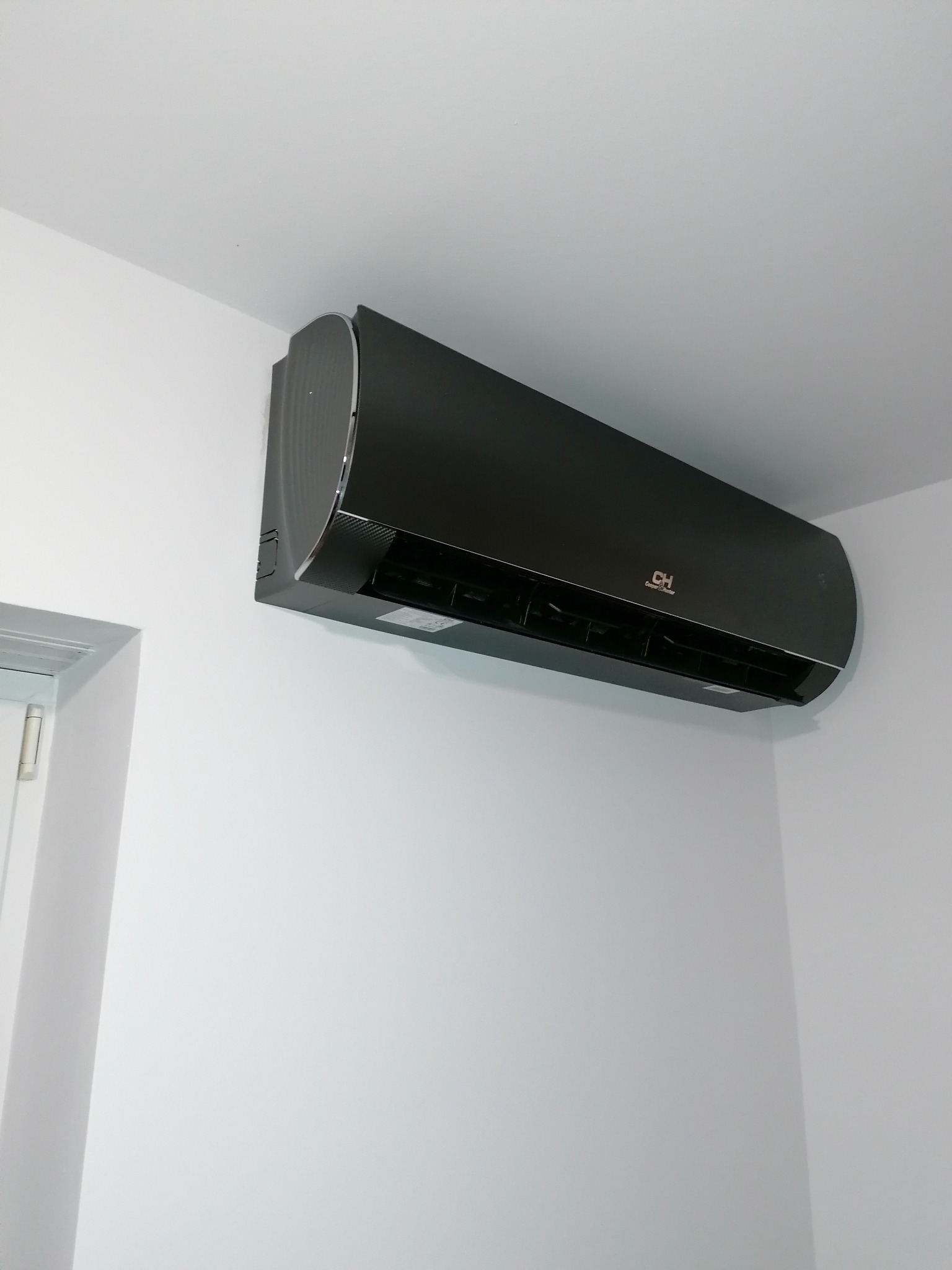 Czarny klimatyzator marki Cooper&Hunter zamontowany na białej ścianie w pomieszczeniu, widoczny fragment ościeżnicy drzwi po lewej stronie.