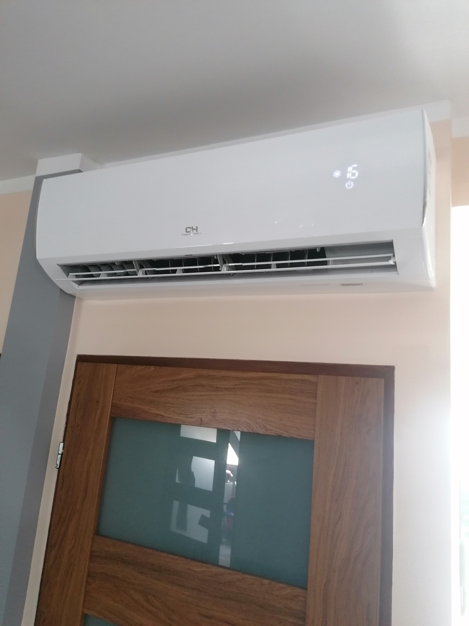 Biały klimatyzator marki CH zamontowany na ścianie nad drewnianymi drzwiami z szybami, widoczny wyświetlacz temperatury.