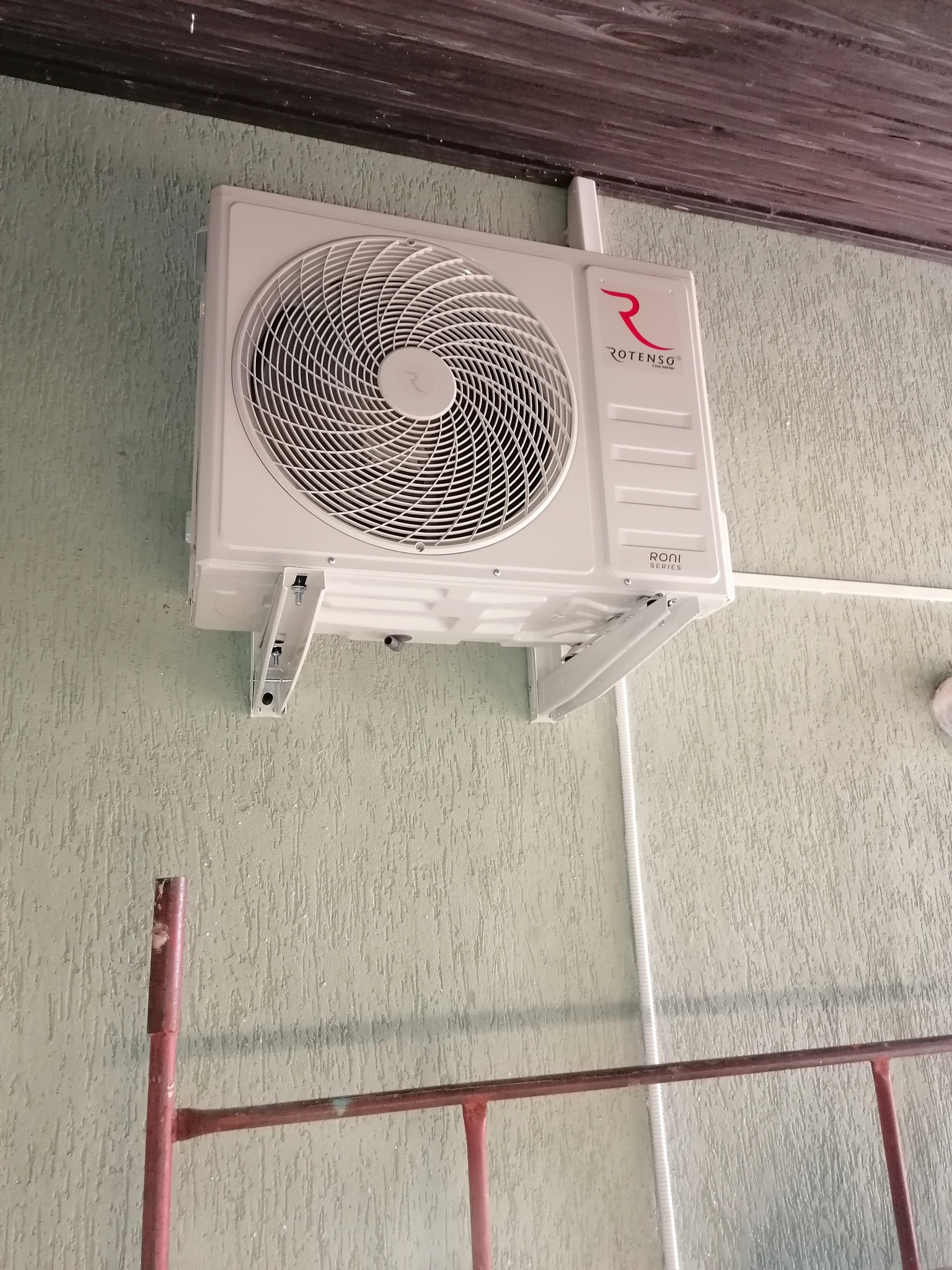 Biała jednostka klimatyzacji Rotenso Roni Series zamontowana na zielonej, teksturowanej ścianie, widoczne metalowe wsporniki i fragment rusztowania malowanego na czerwono.