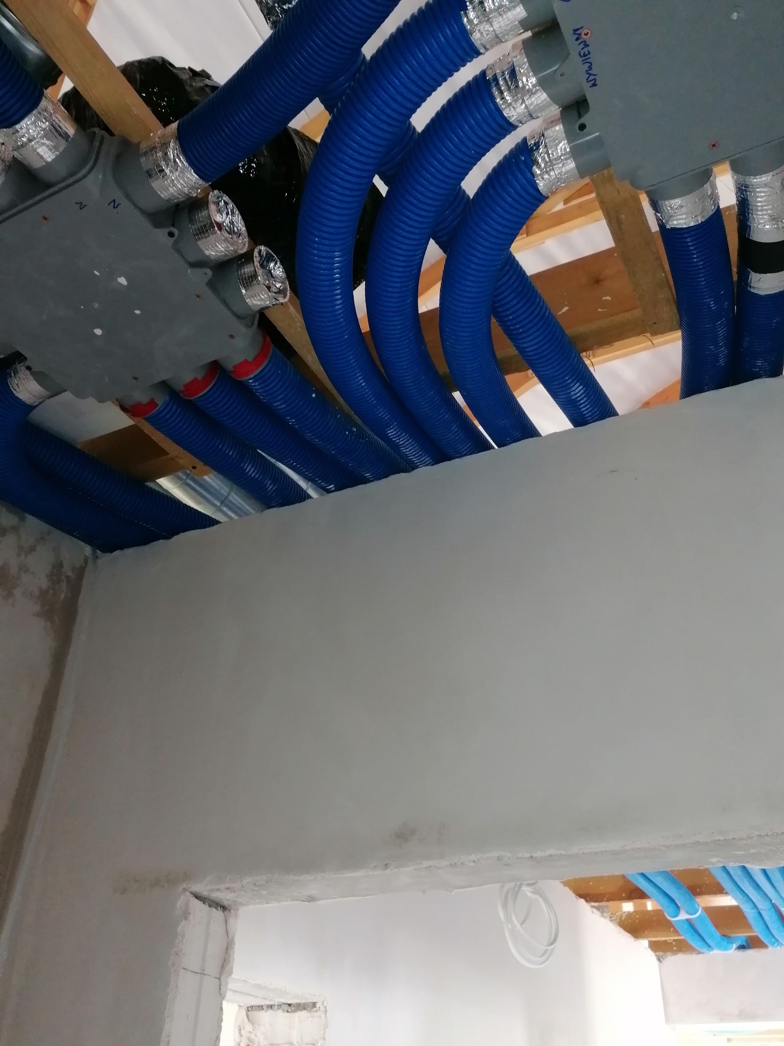 Instalacja systemu rekuperacji z niebieskimi rurami karbowanymi i szarymi rozdzielaczami pod sufitem w trakcie montażu, widoczne elementy konstrukcji drewnianej stropu.