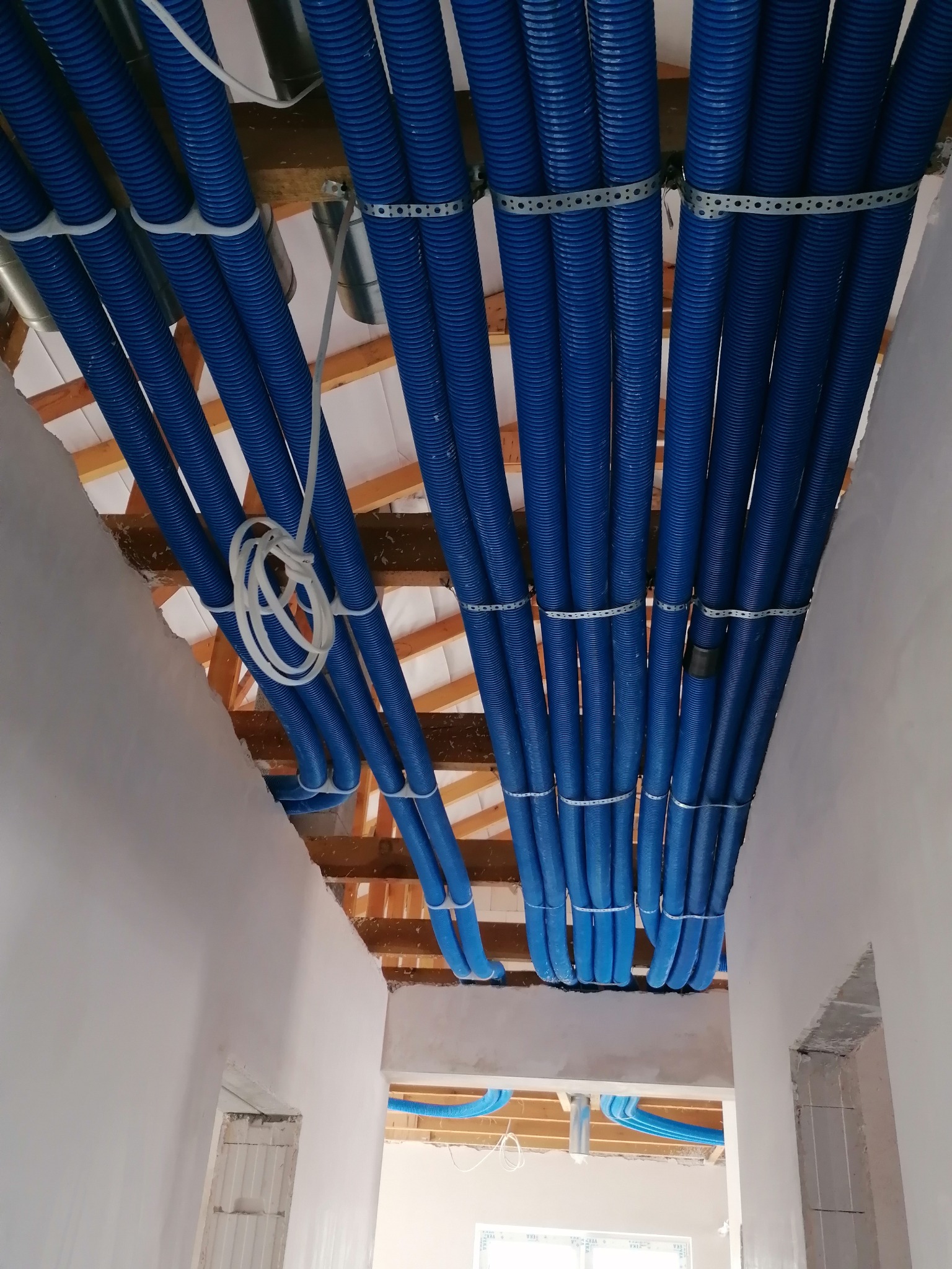 Niebieskie, karbowane rury wentylacyjne zamocowane pod sufitem na drewnianej konstrukcji, spięte metalowymi i plastikowymi obejmami, widok z dołu w surowym wnętrzu.