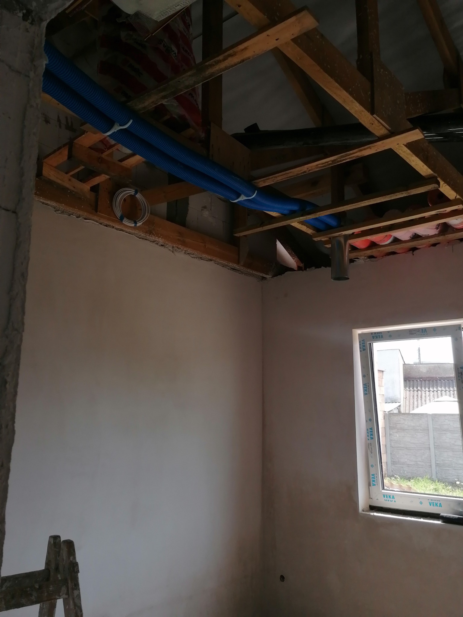 Montaż instalacji wentylacyjnej: niebieskie rury wentylacyjne zamocowane pod drewnianą konstrukcją stropu, widoczne okno i fragment drabiny.