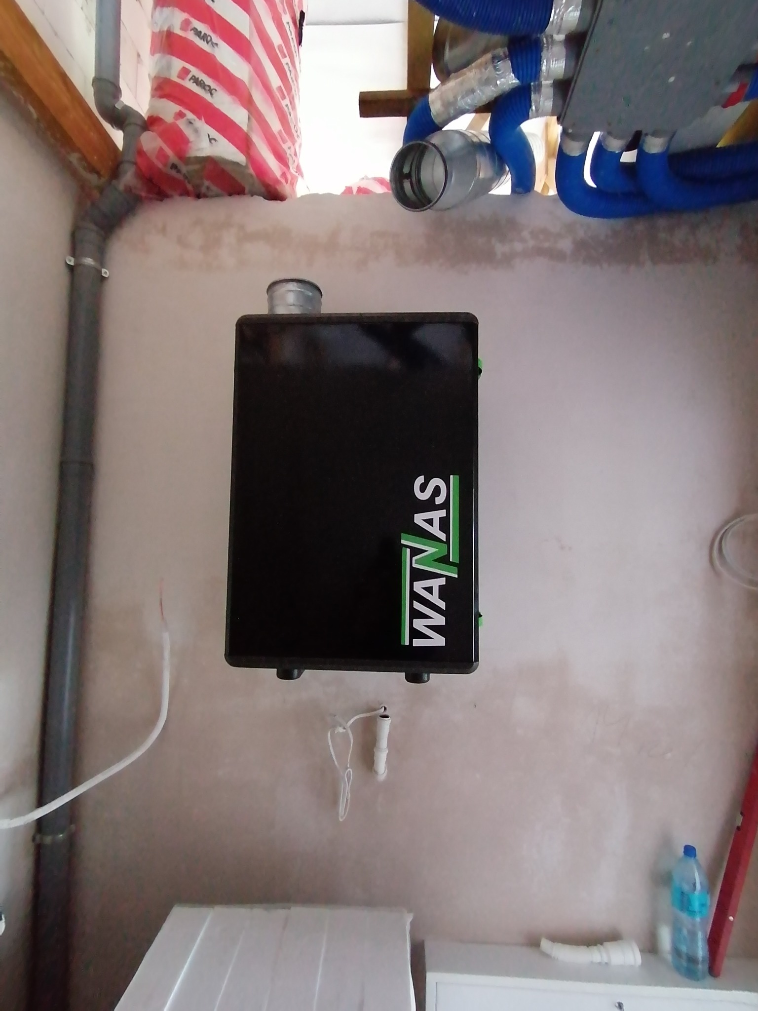 Czarna skrzynka rekuperatora WANAS zamontowana na ścianie w pomieszczeniu w trakcie instalacji, widoczne rury wentylacyjne i izolacja stropu.