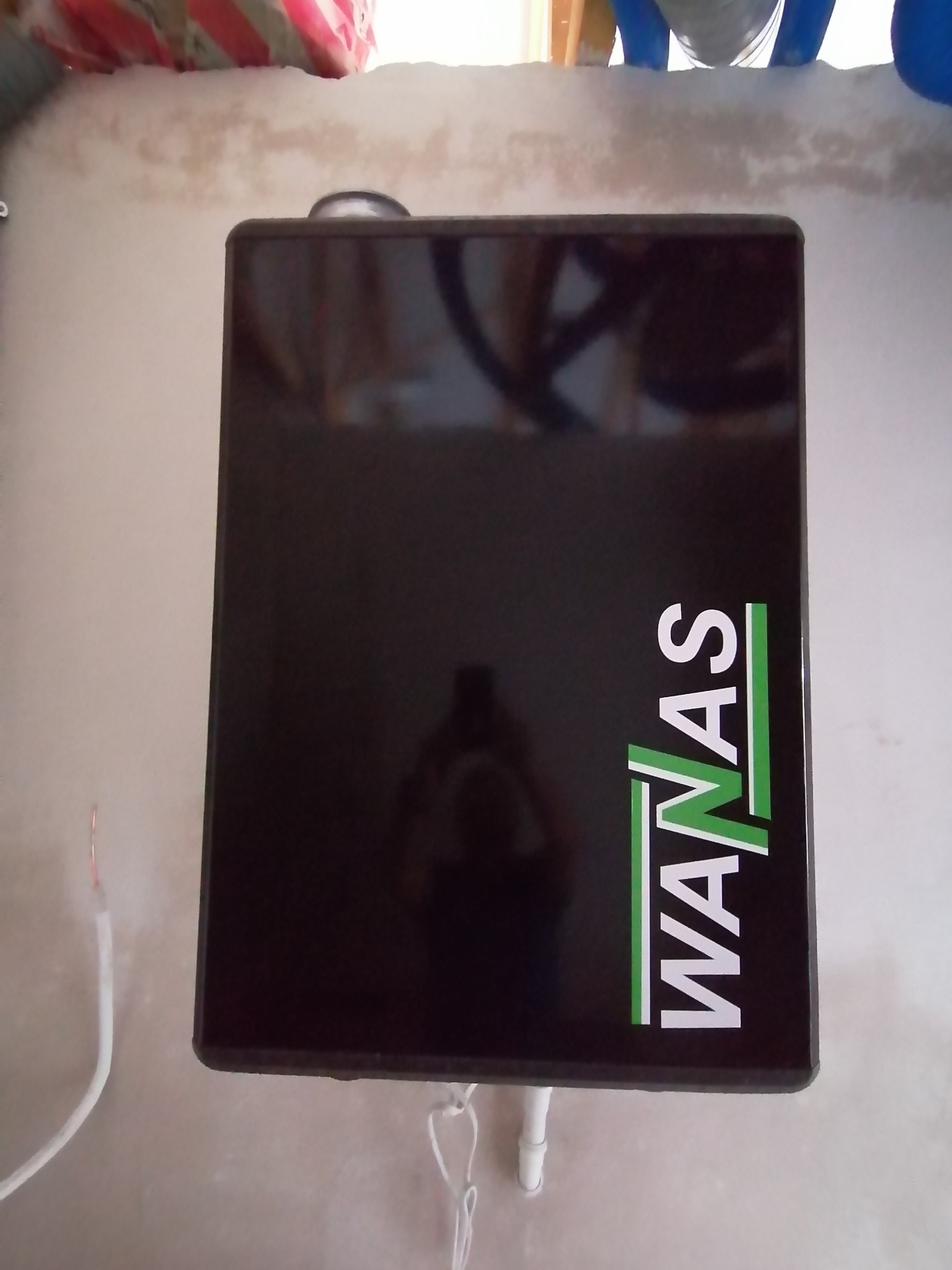 Czarna skrzynka rekuperatora z logo WANAS zamontowana na ścianie, widoczne przewody elektryczne i rurki instalacyjne.
