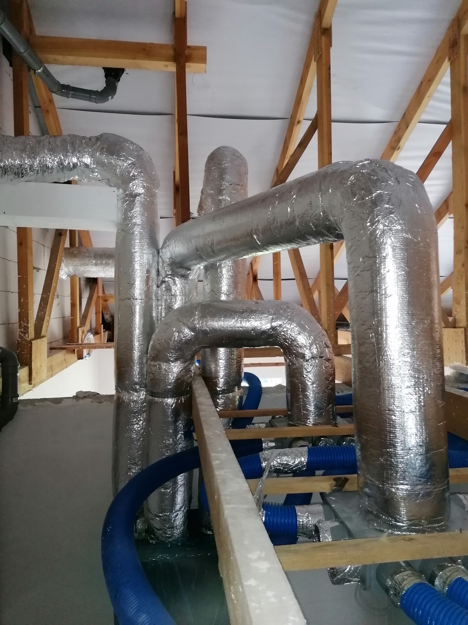 Instalacja rekuperacji z rurami w izolacji aluminiowej i niebieskimi rurami wentylacyjnymi w domu w Kościanie, widok z dołu na drewnianą konstrukcję dachu.