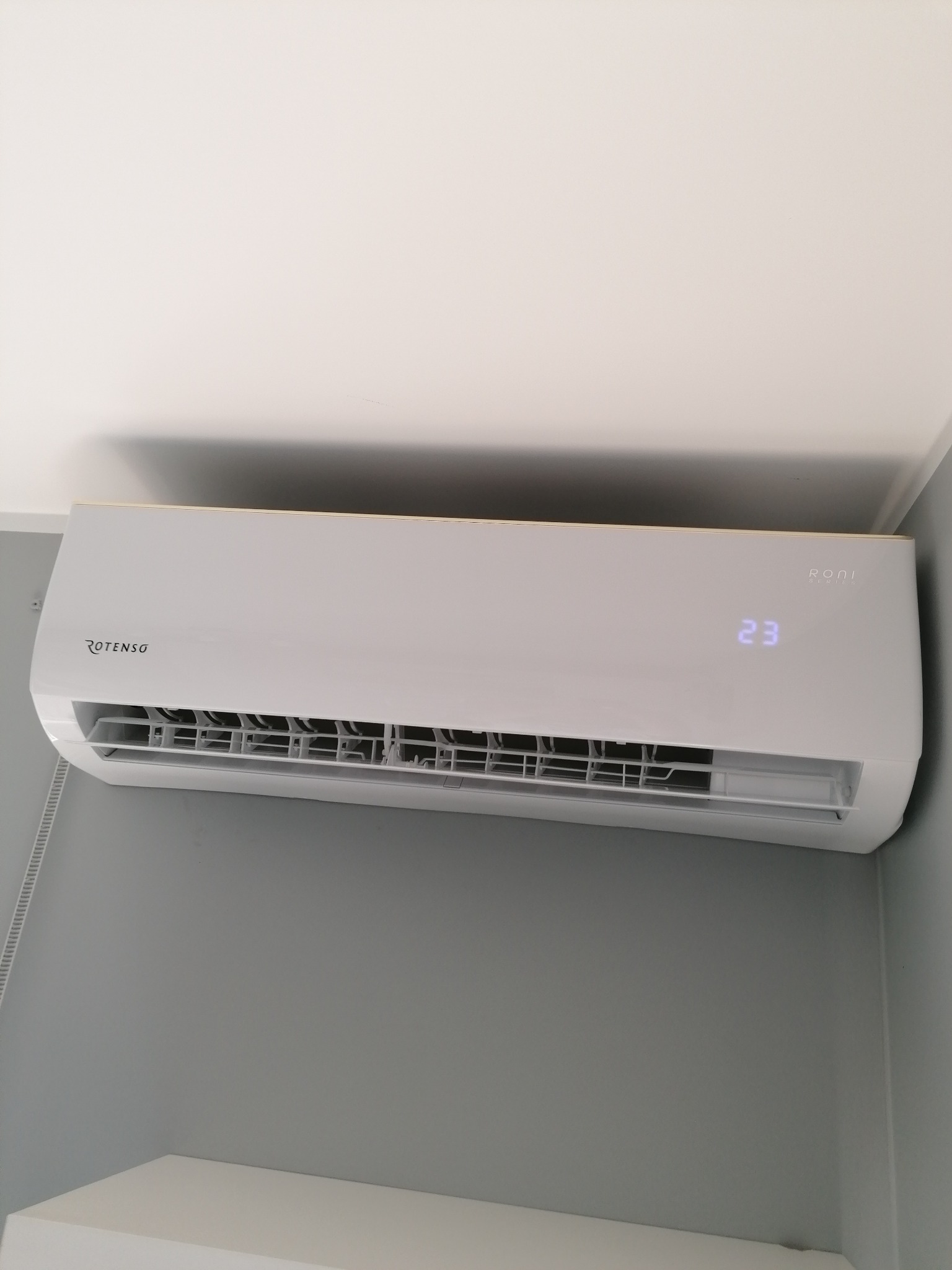 Biały klimatyzator Rotenso Roni Series zamontowany na szarej ścianie, wyświetlający temperaturę 23 stopnie, widoczny fragment białego sufitu i półki.