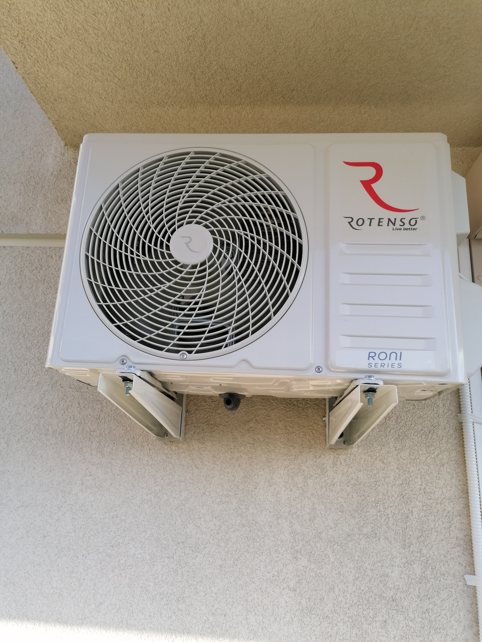 Biała jednostka zewnętrzna klimatyzatora Rotenso Roni Series zamontowana na elewacji budynku, widoczne wsporniki montażowe i peszel z przewodami.
