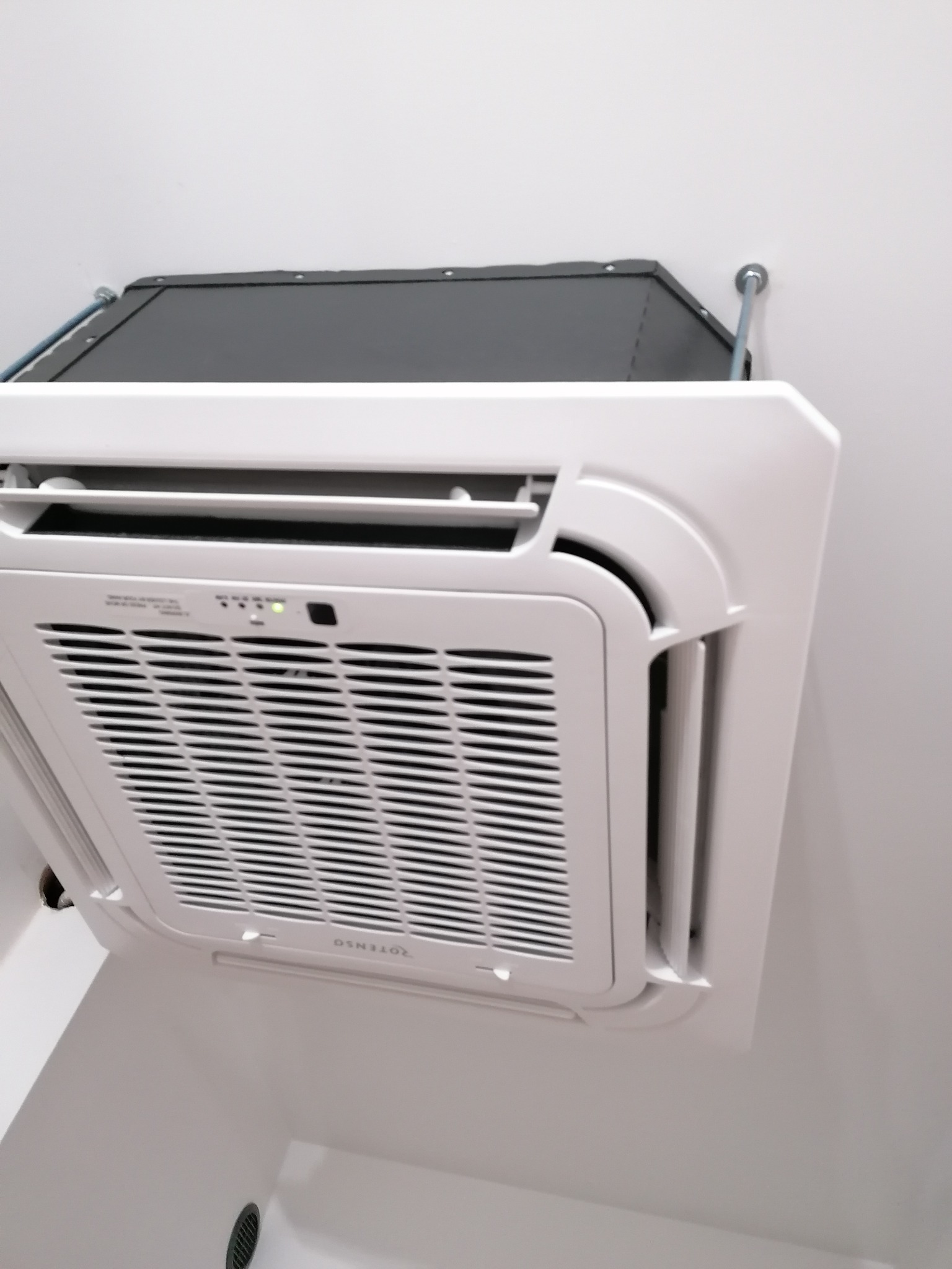 Kasetonowa jednostka klimatyzacji sufitowej marki Rotenso, zamontowana na białym suficie, widoczne diody sygnalizacyjne i elementy montażowe.