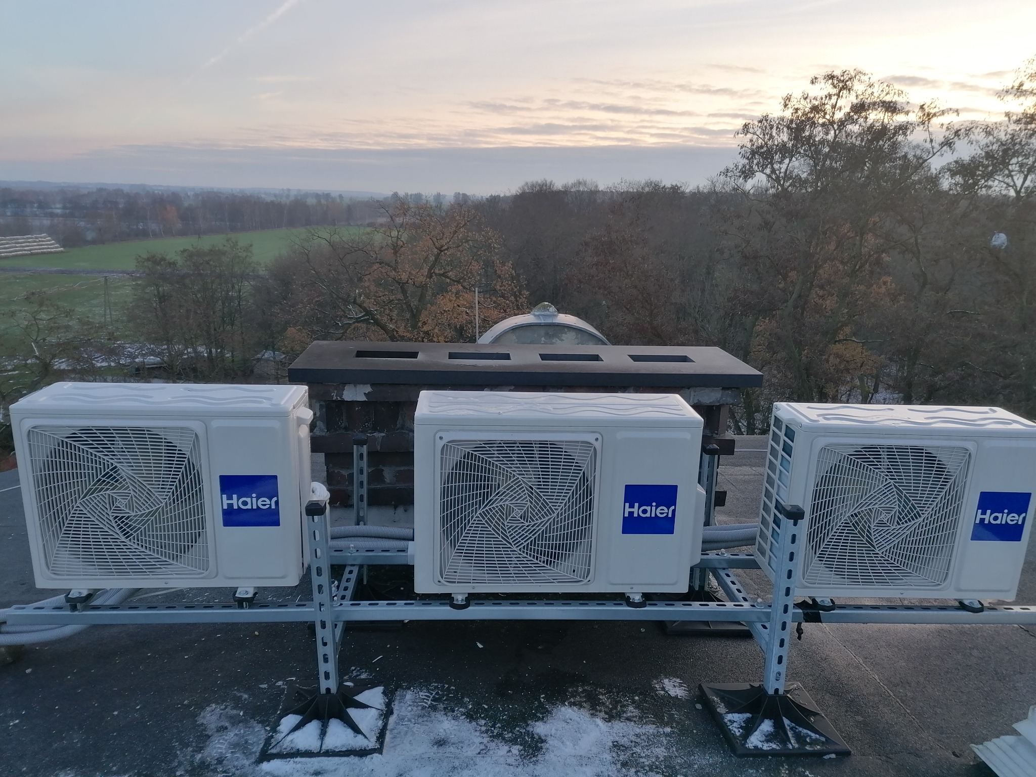 Trzy zewnętrzne jednostki klimatyzacyjne marki Haier zamontowane na dachu budynku, z widokiem na zalesiony krajobraz w tle.
