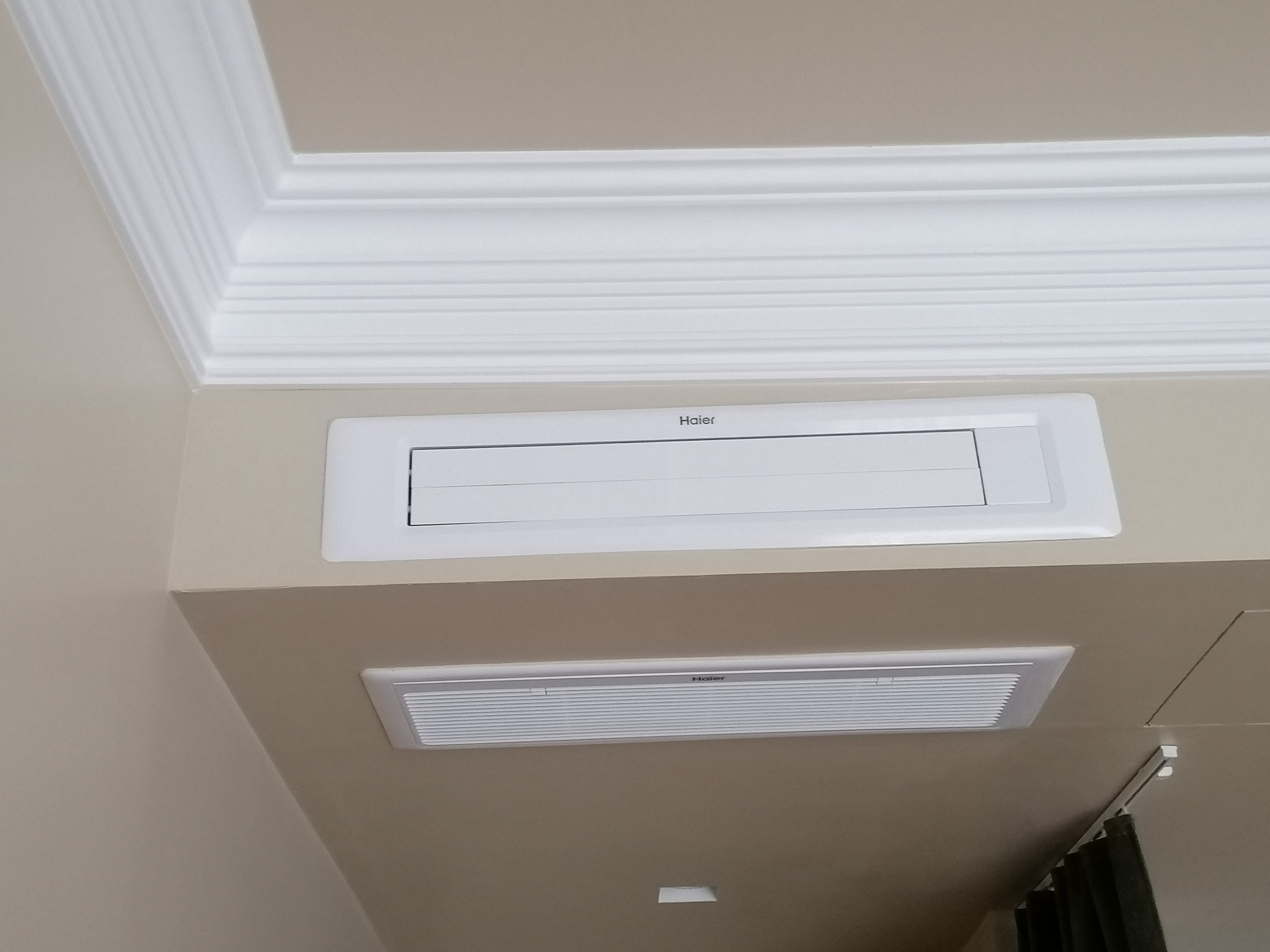 Dwa białe, sufitowe panele klimatyzacyjne marki Haier zamontowane w pomieszczeniu z kremowymi ścianami i ozdobnym gzymsem sufitowym.