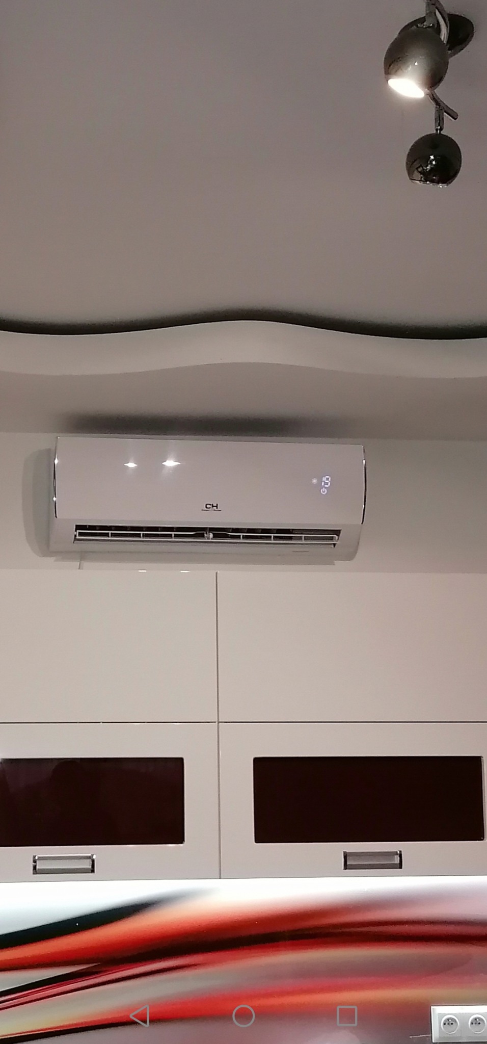 Biała klimatyzacja marki CH zamontowana na ścianie nad szafkami z ciemnymi wstawkami, oświetlona przez reflektor sufitowy