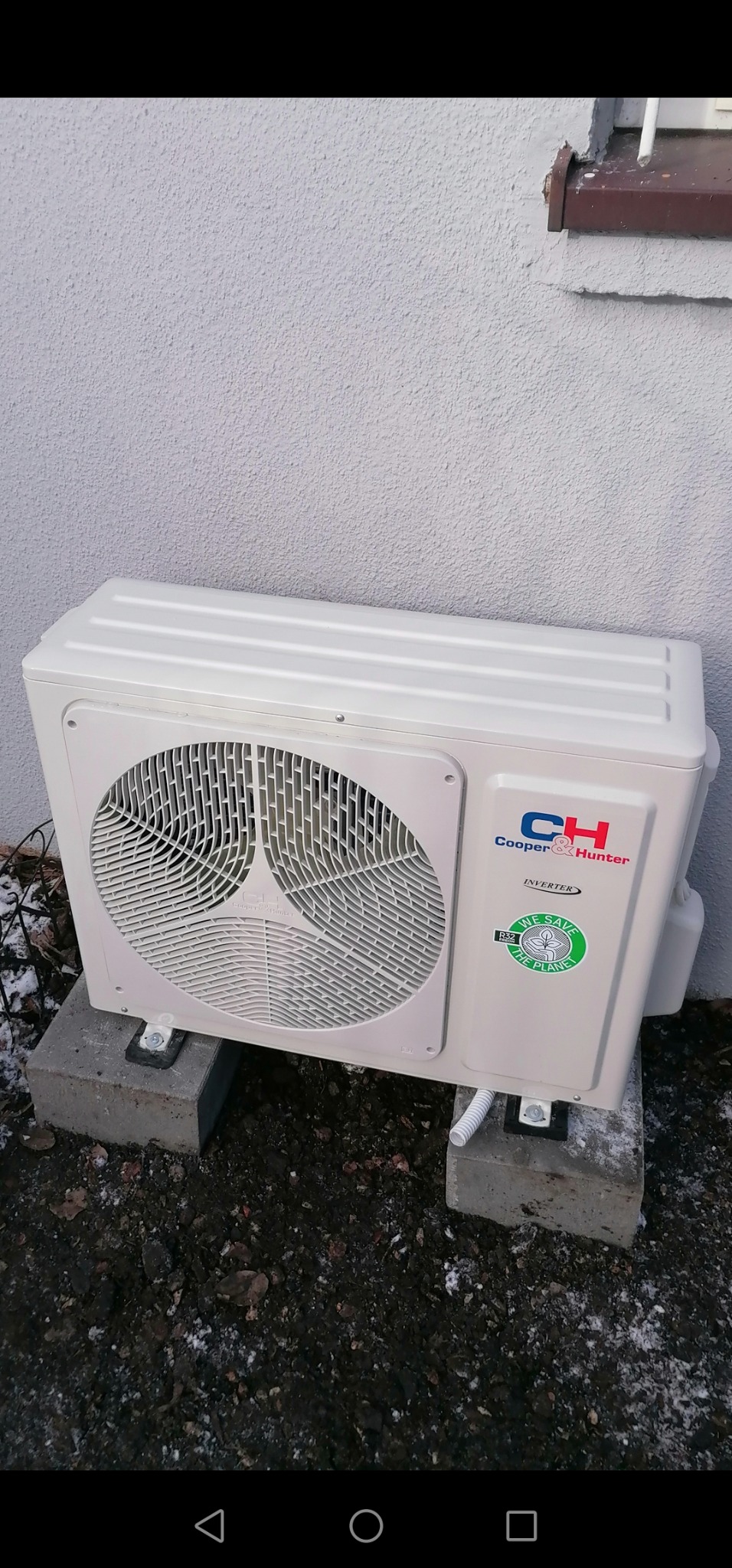 Zewnętrzna jednostka klimatyzacji Cooper&Hunter Inverter, zamontowana na betonowych bloczkach przy ścianie budynku, z widocznym przewodem odprowadzającym skropliny i logotypem 'We save the planet'.