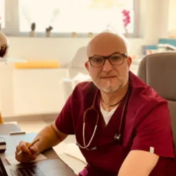 Lekarz w bordowym uniformie i okularach, siedzący przy biurku z laptopem, trzymający długopis nad notatnikiem, w tle okno z kwiatami.