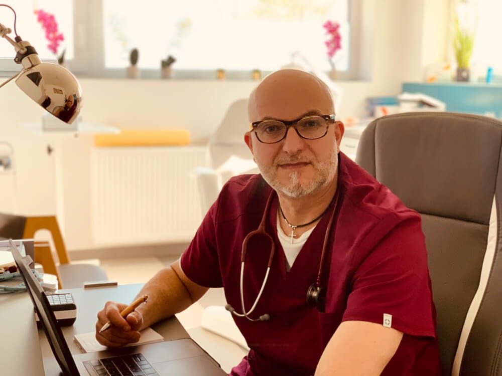 Lekarz w bordowym uniformie i okularach, siedzący przy biurku z laptopem, trzymający długopis nad notatnikiem, w tle okno z kwiatami.