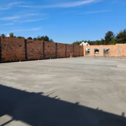 Wylany betonowy fundament pod budynek jednorodzinny, widoczne ściany z czerwonej cegły, wystające rurki instalacyjne i pracownik w odblaskowej kamizelce.