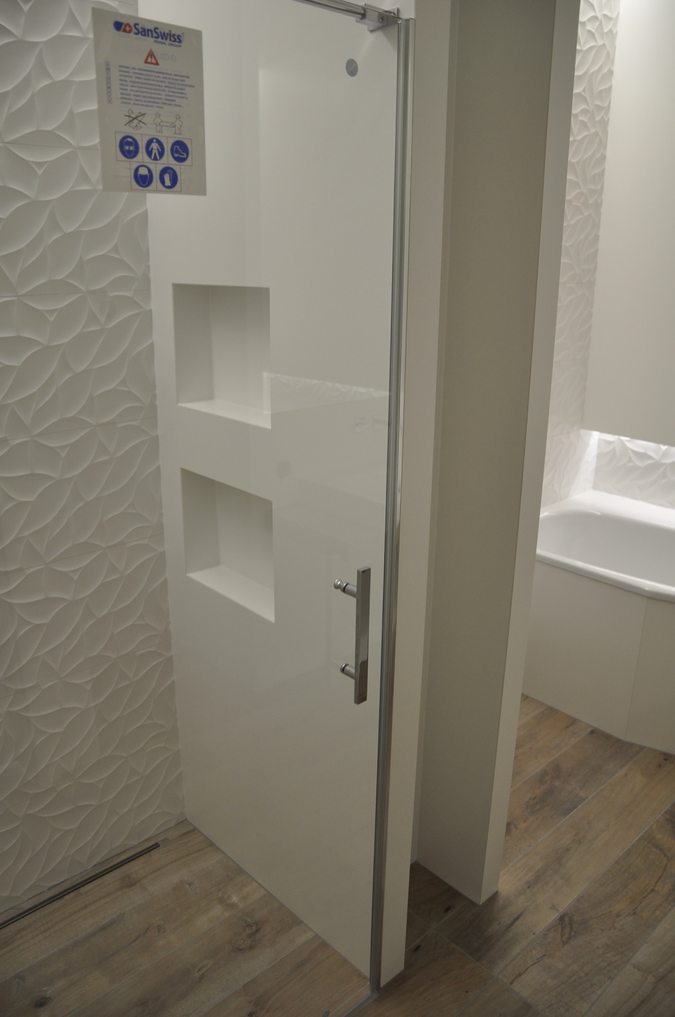 Biała kabina prysznicowa z wnękami w ścianie, wzorzyste płytki na ścianie w tle, drewnopodobna podłoga, widoczna wanna z oświetleniem LED.