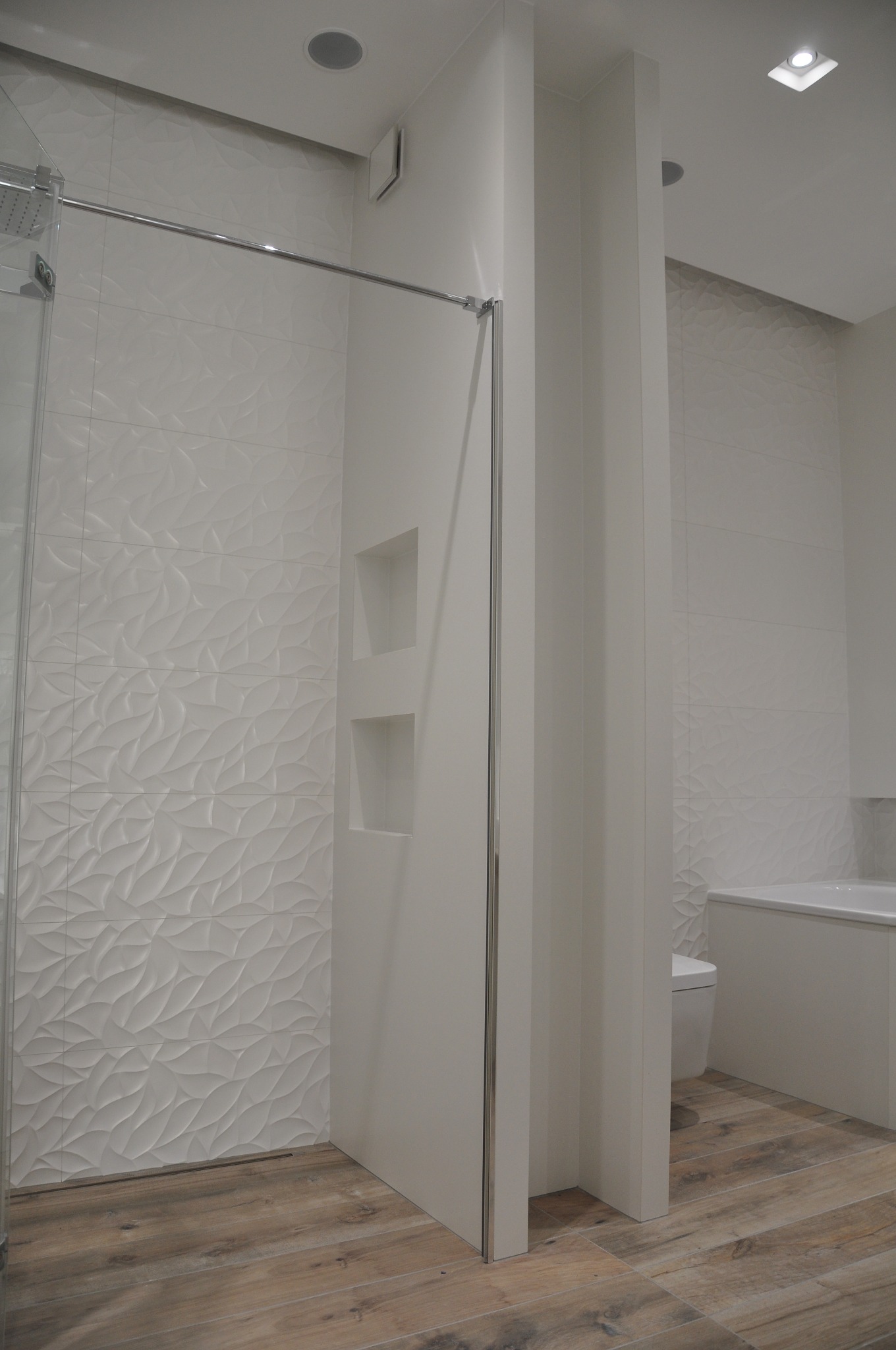 Nowoczesna łazienka z kabiną prysznicową typu walk-in, białe płytki 3D na ścianach, drewnopodobna podłoga, wbudowane półki w ściance działowej.