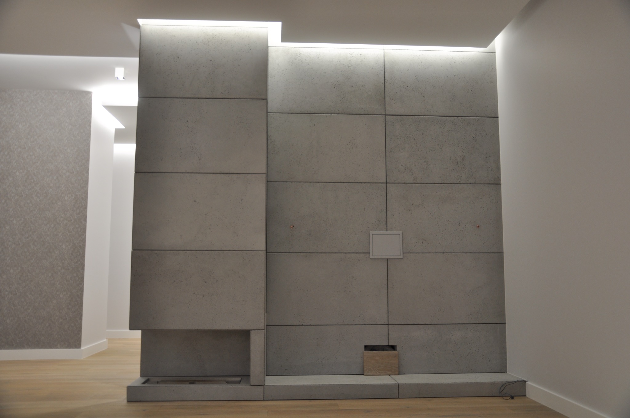 Ściana z szarych, betonowych paneli w nowoczesnym salonie z podświetleniem LED na suficie, mały schowek na drewno i kwadratowa kratka wentylacyjna