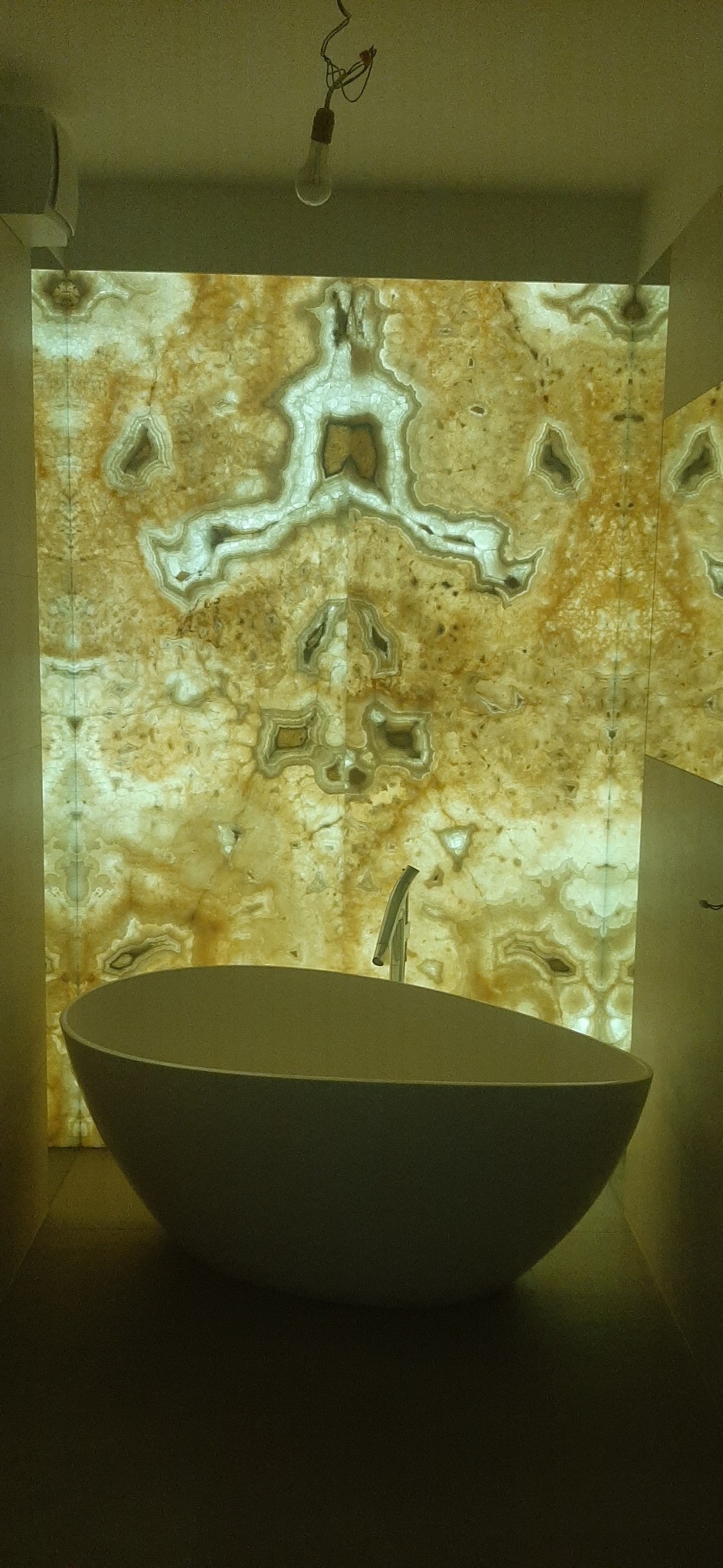 Owalna, wolnostojąca wanna z chromowaną baterią na tle podświetlonej ściany z onyksu o unikalnym wzorze przypominającym agat, widoczna żarówka bez klosza na suficie.