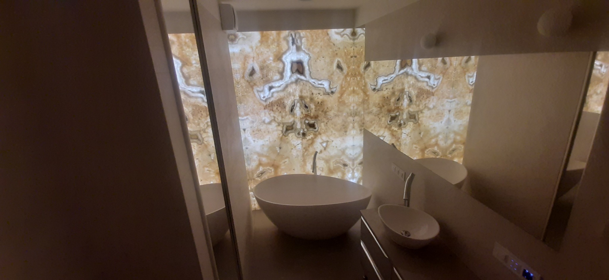 Nowoczesna łazienka z podświetlaną ścianą z onyksu, wolnostojąca umywalka i minimalistyczna armatura.