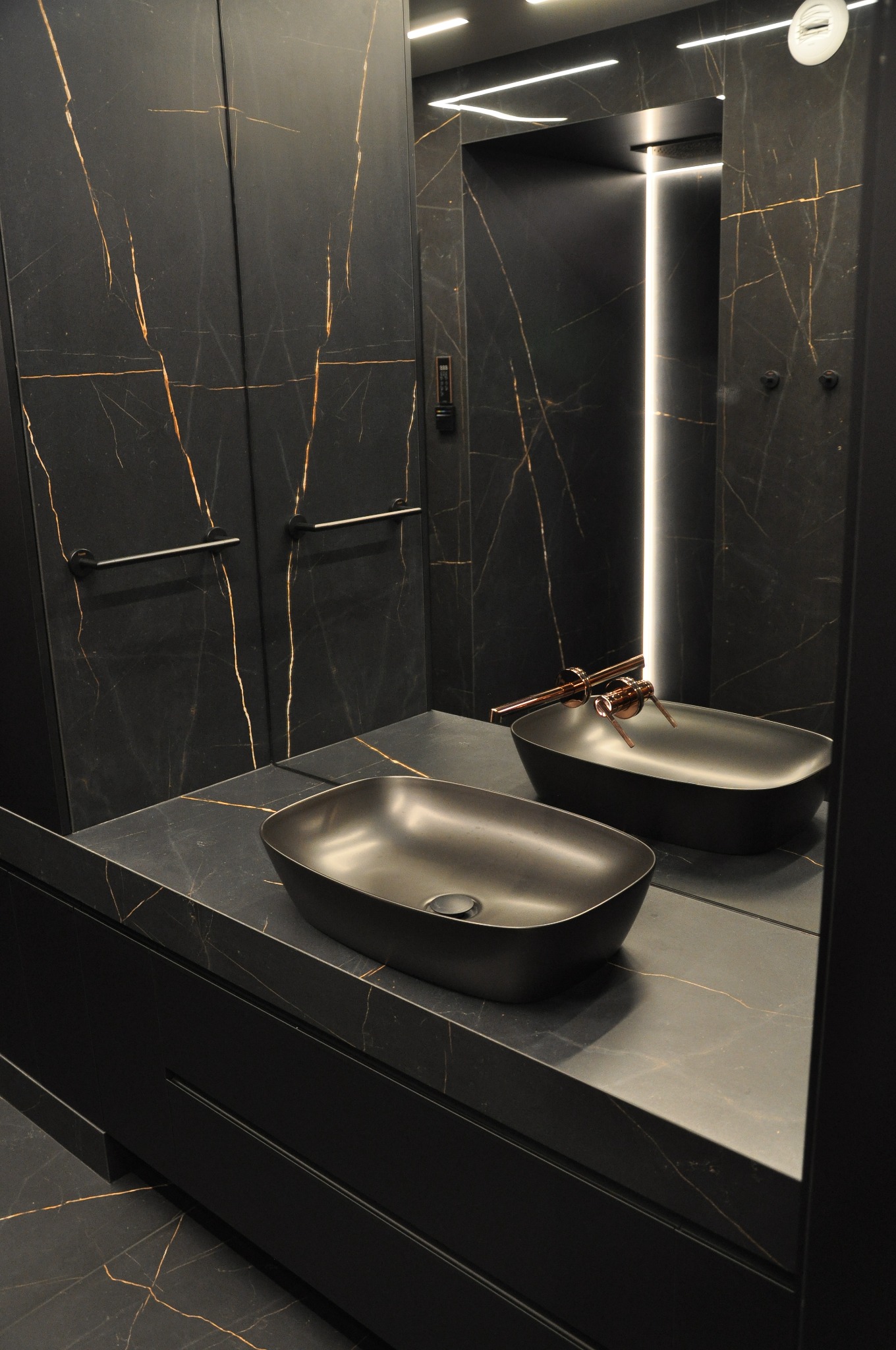 Nowoczesna łazienka z czarnymi marmurowymi ścianami i blatem, dwie matowe, czarne umywalki nablatowe, miedziana bateria, oświetlenie LED w lustrze.