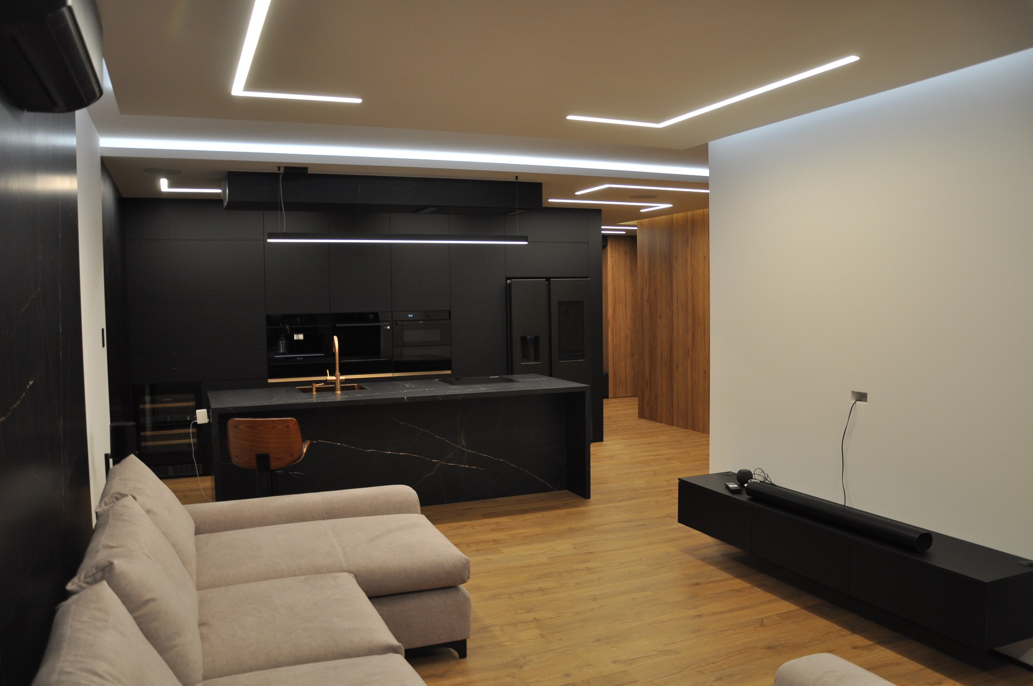 Nowoczesne wnętrze mieszkania: czarna kuchnia z wyspą o marmurowym wzorze, drewniane podłogi i oświetlenie LED, widoczna część sofy w kolorze beżowym oraz szafka RTV w kolorze czarnym.