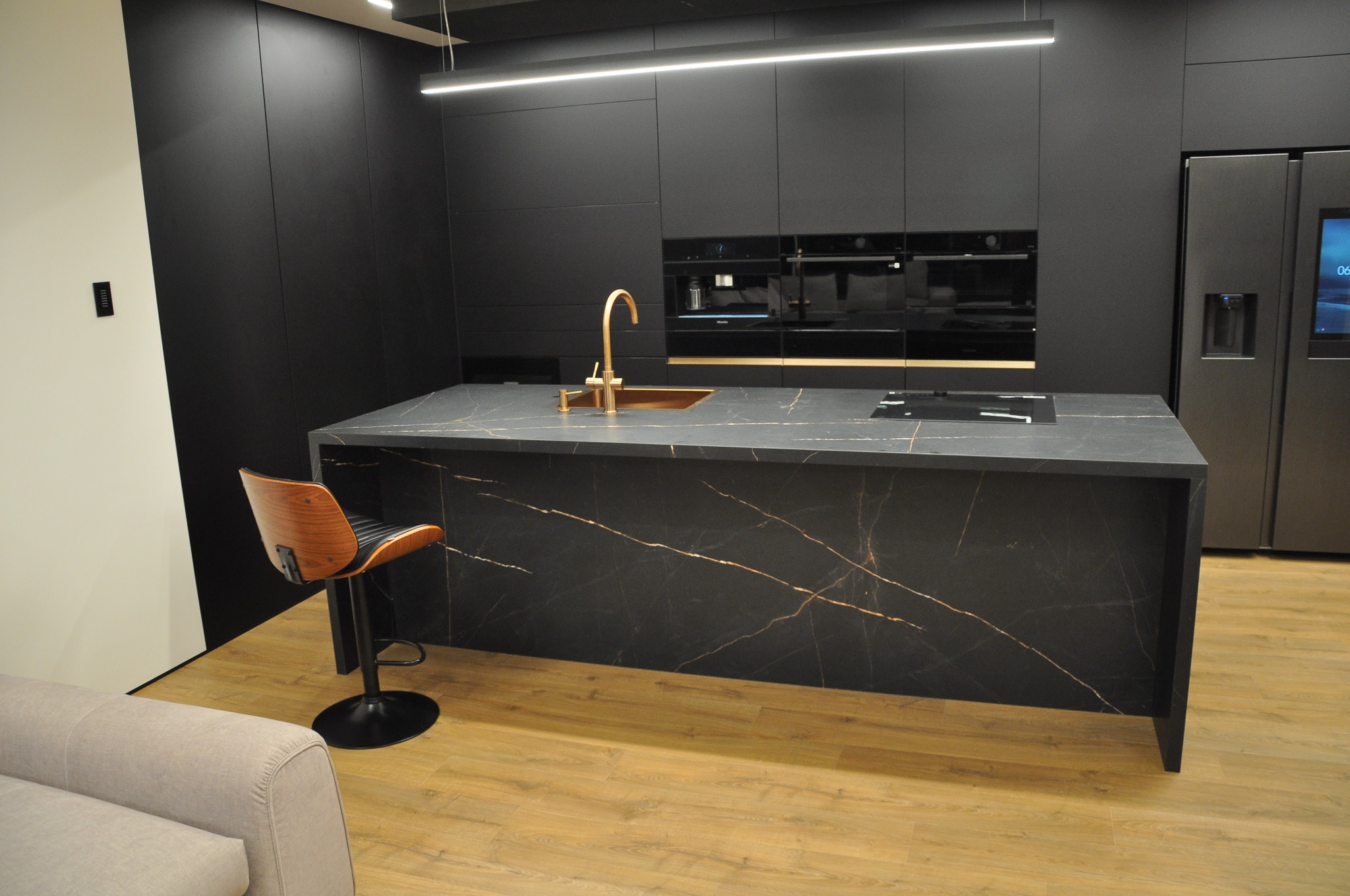 Nowoczesna kuchnia z czarnymi matowymi szafkami, wyspą z blatem imitującym czarny marmur z złotymi żyłami, złotą baterią i hokerem z drewnianym oparciem.