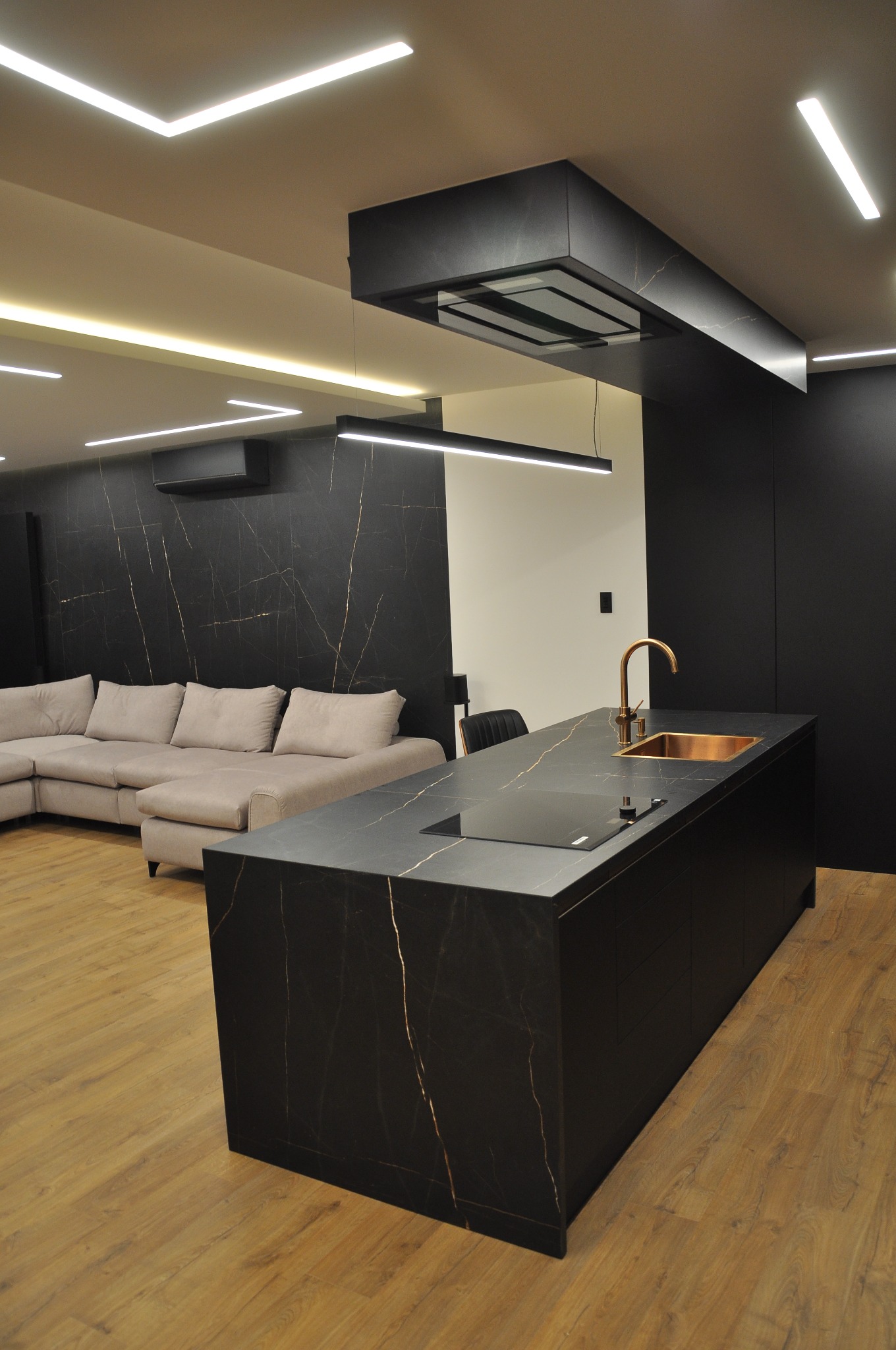 Nowoczesna kuchnia z wyspą wykończoną czarnym marmurem z złotymi żyłami, miedziana bateria i zlew, oświetlenie LED, widoczny salon z szarą sofą narożną.
