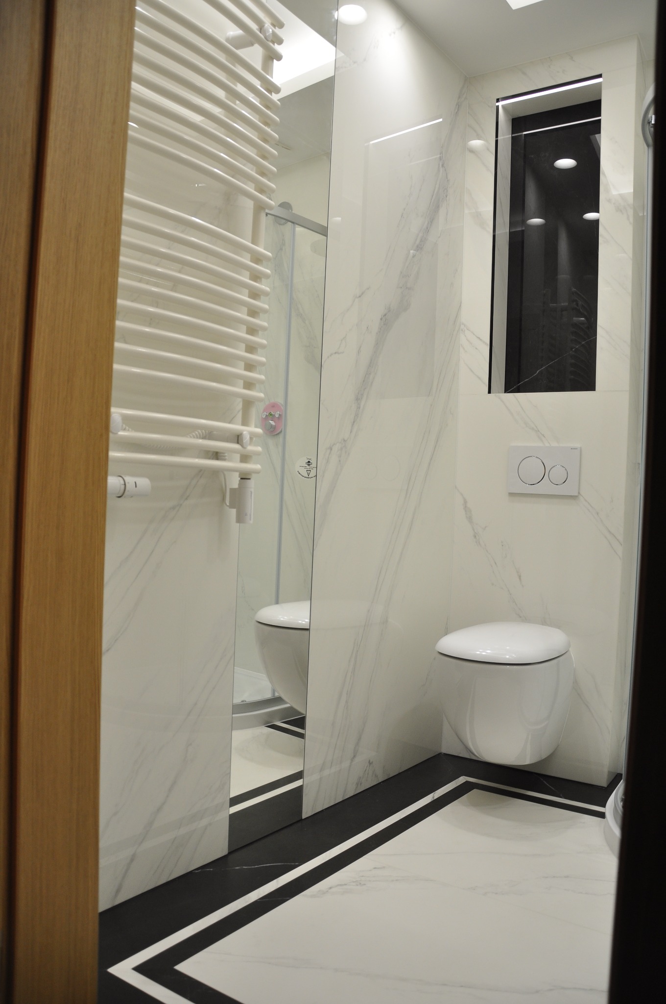 Nowoczesna łazienka z marmurowymi płytkami, podwieszaną toaletą i grzejnikiem drabinkowym, widoczna przez częściowo otwarte, drewniane drzwi.