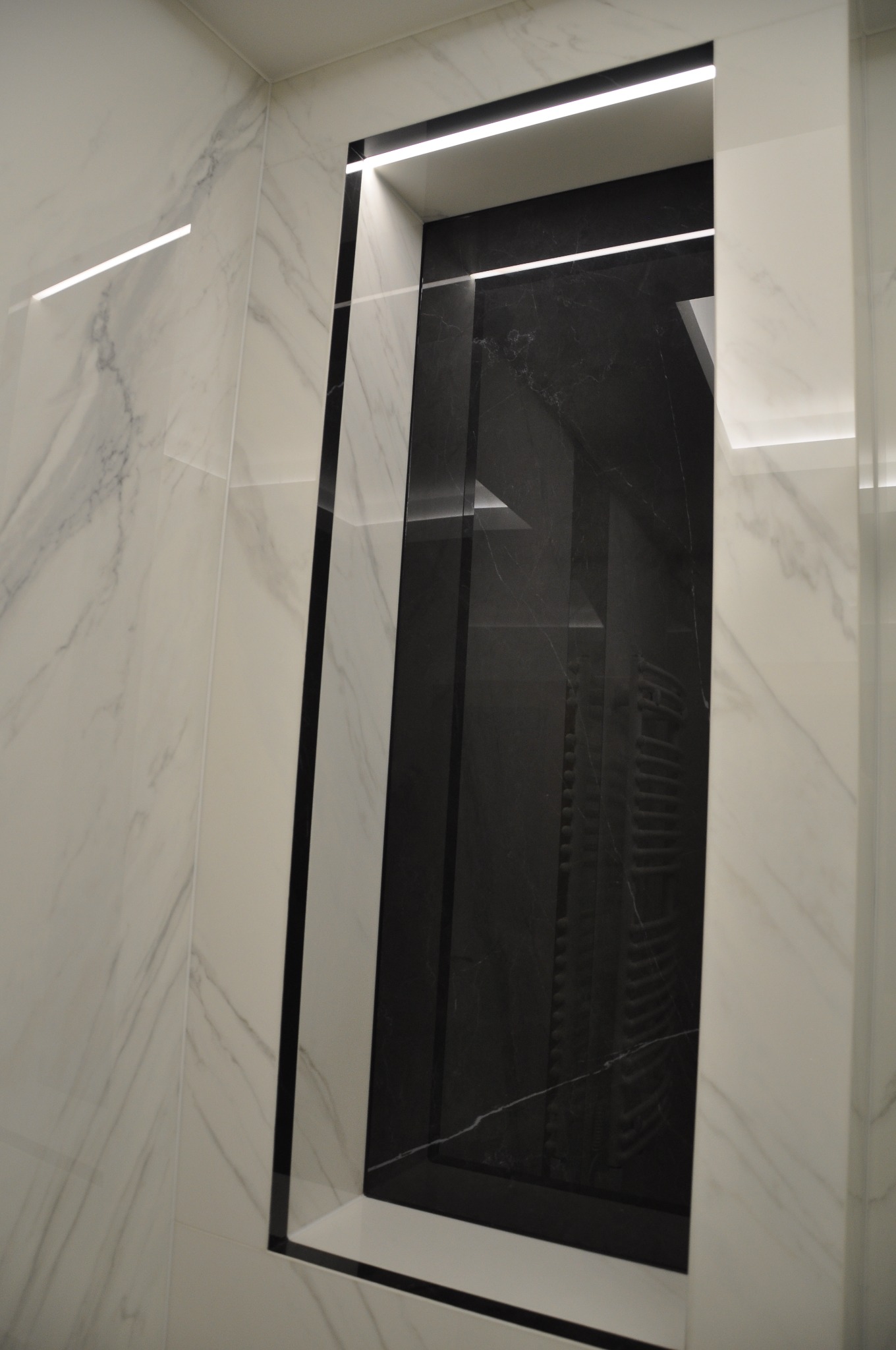 Wnęka w łazience wykończona białym marmurem z szarą żyłą, podświetlona listwą LED, z czarną marmurową ścianą wewnątrz, odbijającą grzejnik.