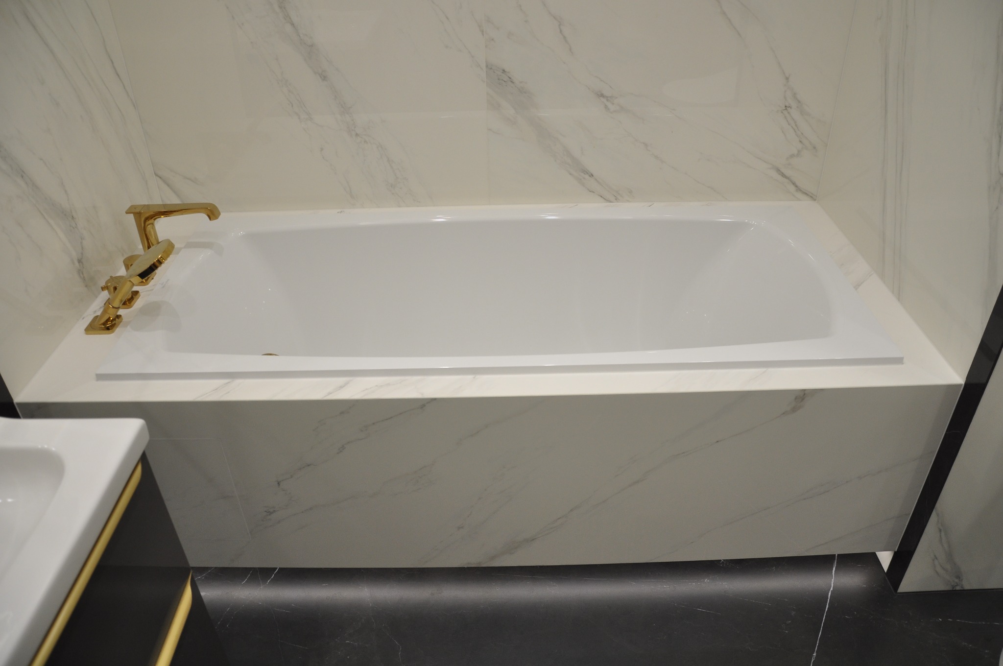 Biała wanna obudowana marmurową płytą, z złotą armaturą na tle marmurowych płytek ściennych, widoczny fragment umywalki.